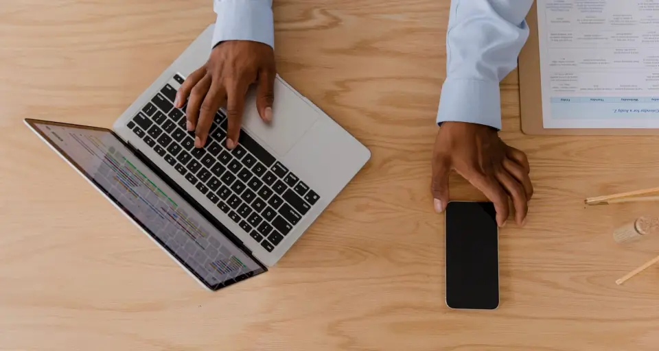 Hands typing on a laptop at a wooden desk, next to a printed page and a phone.